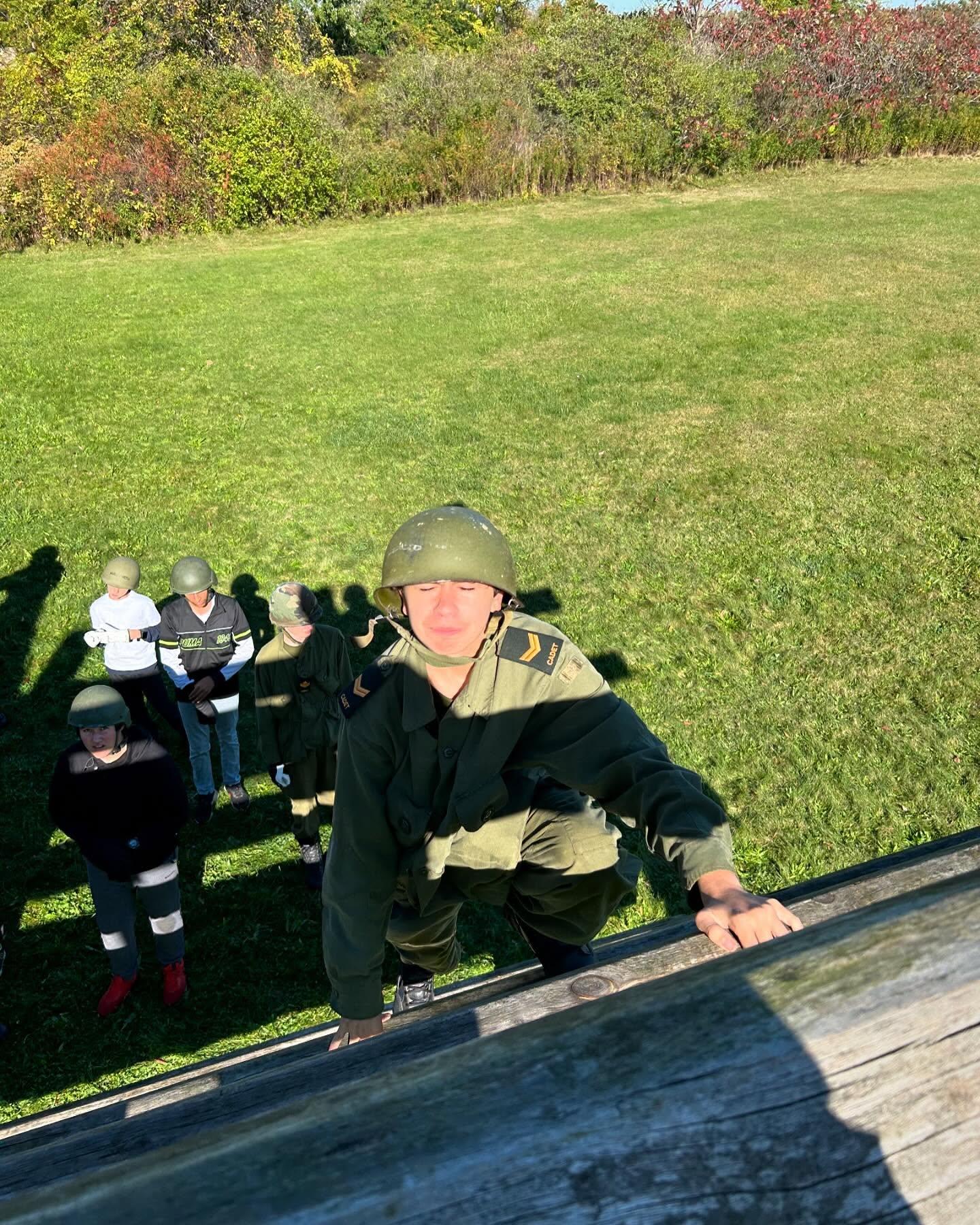 Cadet scaling the wooden obstacle with other cadets gathered below